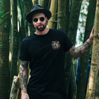 A man standing in a forest of bamboo, wearing the Giant Leap tee, which is a black T-Shirt with an astronaut design on the left breast. The man has tattoos and is wearing sunglasses and a fedora-style hat.